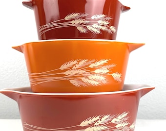 May include: A stack of three vintage Pyrex casserole dishes in shades of red and orange. Each dish features a wheat stalk design in white. The dishes have handles and are nested for display.