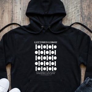 Puede incluir: Una sudadera negra con capucha que presenta un estampado gráfico blanco de Gottfried Leibniz con un patrón repetitivo de círculos y líneas verticales, junto al texto: Every substance is like an entire world and like a mirror of God.