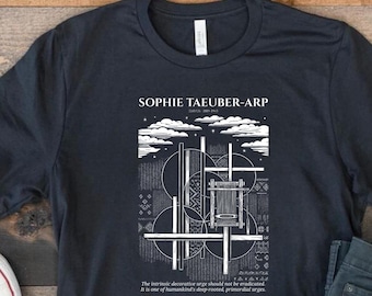 May include: A dark charcoal gray short-sleeve t-shirt featuring a white graphic print of Sophie Taeuber-Arp artwork. The design includes geometric shapes, a loom illustration, clouds, and text that reads, The intrinsic decorative urge should not be eradicated. It is one of humankind's deep-rooted, primordial urges.