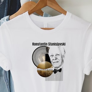 Può includere: T-shirt bianca con grafica di Konstantin Stanislavskij, un ritratto e disegni circolari astratti in nero e oro. Il testo "Konstantin Stanislavskij" è sopra la grafica. La maglietta è appesa a una gruccia di legno.