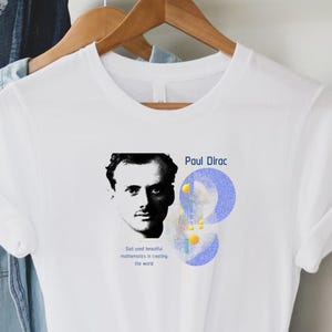 May include: White t-shirt with a black and white portrait of Paul Dirac, his name in blue, and a quote. The design includes blue and yellow abstract shapes.