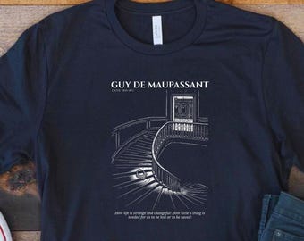 May include: A dark navy blue short-sleeve t-shirt featuring a white line art illustration of a curved staircase. The text reads GUY DE MAUPASSANT and How life is strange and changeful! How little a thing is needed for us to be lost or to be saved!