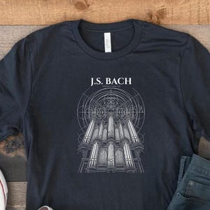 Może przedstawiać: Ciemnoszary t-shirt z krótkim rękawem z białą grafiką liniową przedstawiającą organy piszczałkowe pod napisem J.S. BACH. Koszulka jest prezentowana wraz z parą białych tenisówek z płótna i ciemnoszarymi dżinsami.