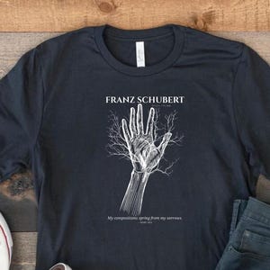 May include: Dark navy blue t-shirt featuring a white graphic of a hand with tree-like veins, and the text "FRANZ SCHUBERT" above and a quote below. The shirt is paired with blue jeans and white canvas sneakers.