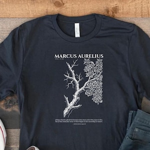 Pode incluir: Uma t-shirt cinzento-escura de manga curta com um gráfico branco de uma árvore que está nua de um lado e florida do outro. O texto diz MARCUS AURELIUS e inclui uma citação sobre viver de acordo com a natureza sob a árvore.