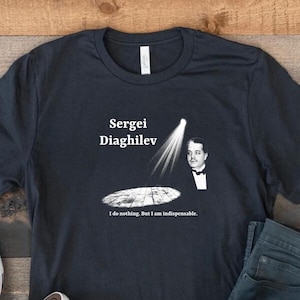 Może przedstawiać: Ciemnoniebieska koszulka z napisem "Sergei Diaghilev" i grafiką mężczyzny w reflektorze. Poniżej grafiki znajduje się cytat "I do nothing. But I am indispensable.". Na zdjęciu znajdują się również białe trampki Converse i niebieskie dżinsy.