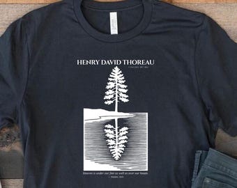 May include: A dark gray short-sleeve t-shirt featuring a white graphic of a pine tree reflected in water. The text reads HENRY DAVID THOREAU and Heaven is under our feet as well as over our heads. A pair of white canvas sneakers and dark jeans.
