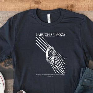May include: A dark charcoal gray short-sleeve t-shirt featuring a white graphic of a ring with radiating lines. The text reads BARUCH SPINOZA and All things excellent are as difficult as they are rare. The shirt is paired with white sneakers and jeans.