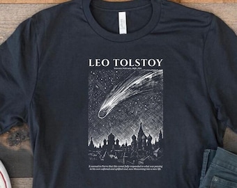 May include: A dark charcoal gray short-sleeve t-shirt featuring a white graphic print of a comet over a city skyline with onion domes. The text reads LEO TOLSTOY and includes a quote below the illustration. Dark gray jeans and white sneakers.