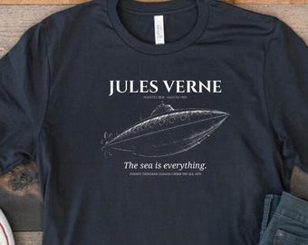 Puede incluir: Una camiseta azul marino con el texto "JULES VERNE" y una ilustración blanca de un submarino. La frase "The sea is everything" está impresa debajo. La camiseta se combina con vaqueros azules y zapatillas blancas.