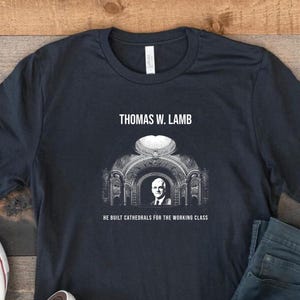 May include: Dark navy blue t-shirt featuring a graphic of Thomas W. Lamb, an architectural rendering, and the text "He Built Cathedrals for the Working Class." The shirt is paired with blue jeans and white canvas sneakers.