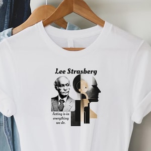 Può includere: T-shirt bianco con un ritratto in bianco e nero di Lee Strasberg, la scritta "Lee Strasberg" sopra e "Acting is in everything we do." sotto. La maglietta presenta anche una grafica di due volti.