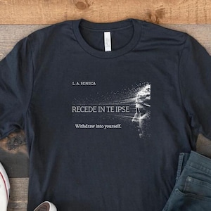 May include: Dark navy blue t-shirt with the text "RECEDE IN TE IPSE" and "Withdraw into yourself." A pair of white sneakers and blue jeans are next to the shirt. The shirt also has the text "L.A. SENECA".