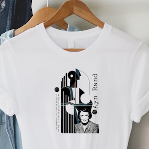 以下が含まれることがあります： 「Ayn Rand」の文字と白黒のグラフィックデザインが特徴の白いTシャツ。デザインには肖像画と抽象的な形が含まれています。木製のハンガーにTシャツが掛けられています。