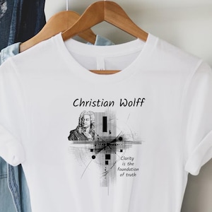 May include: White t-shirt featuring the name "Christian Wolff" above a black and white portrait and abstract design. The text "Clarity is the foundation of truth" is below the design. The shirt is hanging on a wooden hanger.