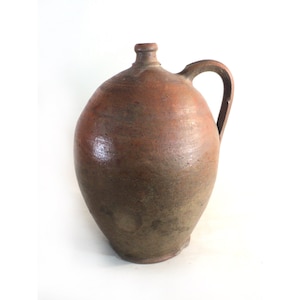 May include: A large, brown, earthenware jug with a single handle. The jug has a rounded body and a slightly flared lip.