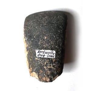 以下が含まれることがあります： ざらざらした質感と丸みを帯びた形状の濃い灰色の石。 「Bordouville drag-Seine」と書かれた黒い文字の小さな白いラベルが石に貼られています。 石は白い背景に配置されています。