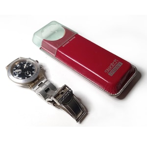 May include: A Swatch Irony watch with a black face and silver band, next to its red and clear case. The watch case is rectangular with the words "Swatch Irony" printed on it. The watch is Swiss made.