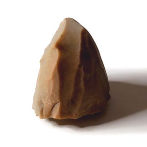 以下が含まれることがあります： 明るい茶色の、ほぼ三角形の石器で、表面はざらざらしています。石は鋭いエッジと丸みを帯びた上部を持ち、色の層が見えます。白い背景に置かれ、影を落としています。