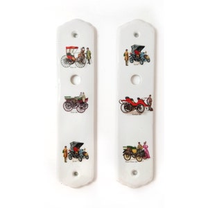Op de afbeelding: Twee witte keramische wandplaten met illustraties van vintage auto's en mensen. Elke plaat heeft drie gaten en een decoratieve bovenkant. De auto's zijn in verschillende kleuren, met mensen in kleding uit die tijd.