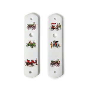 May include: Two white ceramic door pulls featuring images of vintage cars. Each pull showcases three different cars, including a red car, a green car, and a black car.