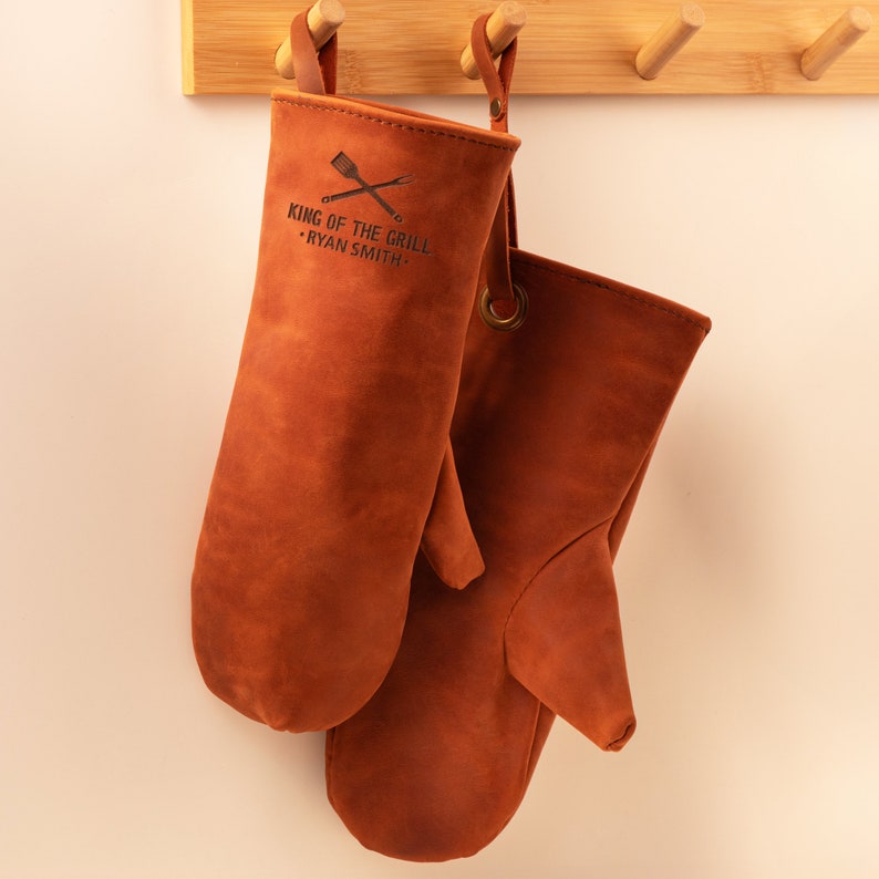 May include: Two brown leather oven mitts hanging on a wooden rack. The left mitt has the text "King of the Grill - Ryan Smith" embossed on it.