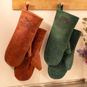 May include: Two leather oven mitts hanging on a wooden rack. The brown mitt has the text "King of the Grill - Ryan Smith" and the green mitt has the text "Amelia's Kitchen".