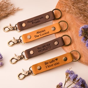 May include: Four rectangular leather keychains with bronze-toned metal lobster clasps and key rings. Two are dark brown and two are tan. Each features a metal rivet and laser-engraved text: Please drive safe Kaitlin, Always The Coolest, You are our WORLD, and Be safe I love you.