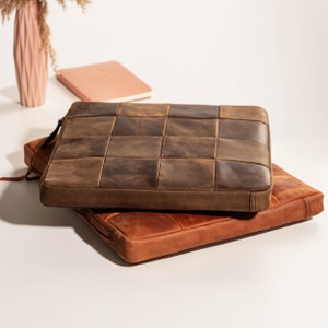 May include: Two leather laptop sleeves stacked on top of each other. The top sleeve is brown with a patchwork design. The bottom sleeve is a lighter brown color.