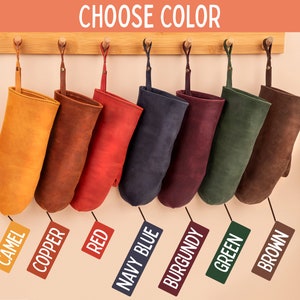 May include: Eight oven mitts hanging on a wooden rack, each in a different color: camel, copper, red, navy blue, burgundy, green, and brown. The mitts are made of leather and have a rustic look. "Choose Color" is written at the top of the image.