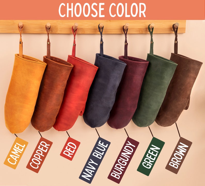 May include: Eight oven mitts hanging on a wooden rack, each in a different color: camel, copper, red, navy blue, burgundy, green, and brown. The mitts are made of leather and have a rustic look. "Choose Color" is written at the top of the image.