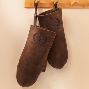 May include: Two brown leather oven mitts with a black embroidered logo that says "Real Grill Master" hanging on a wooden rack.