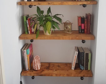 May include: Four wooden shelves with black metal brackets, holding books, a potted plant, a small clock, and a decorative salt lamp. The shelves are mounted on a light blue wall.