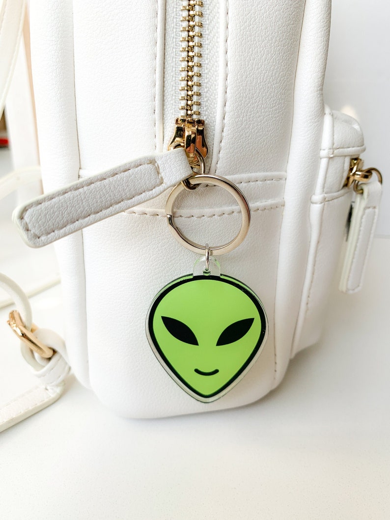 May include: A white backpack with a green alien face keychain attached to the zipper pull. The keychain is made of clear acrylic with a black outline.