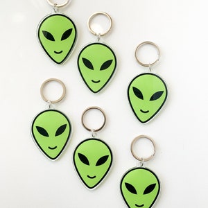 May include: Six green alien face keychains with silver key rings. The alien faces have black eyes and a smiling mouth.