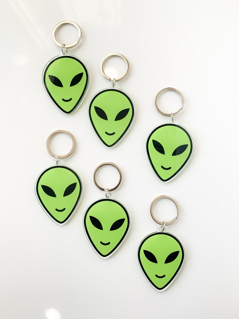 May include: Six green alien face keychains with silver key rings. The alien faces have black eyes and a smiling mouth.