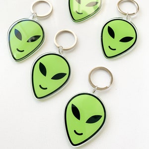 May include: Six green alien face keychains with silver key rings. The alien faces are smiling with black eyes and a black outline.