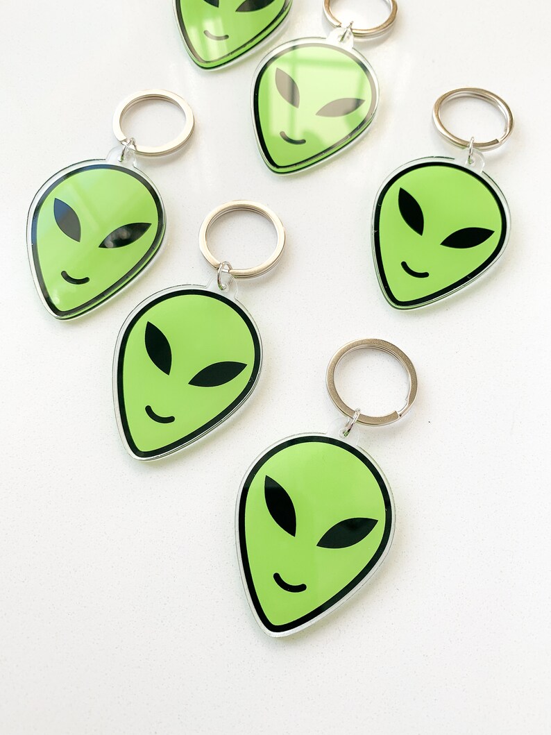 May include: Six green alien face keychains with silver key rings. The alien faces are smiling with black eyes and a black outline.