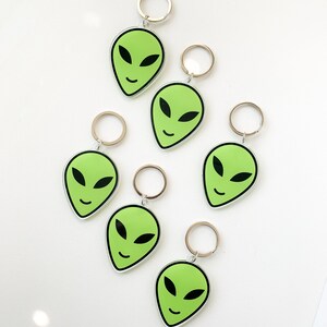 May include: Six green alien face keychains with black eyes and a smile. Each keychain has a silver metal ring.
