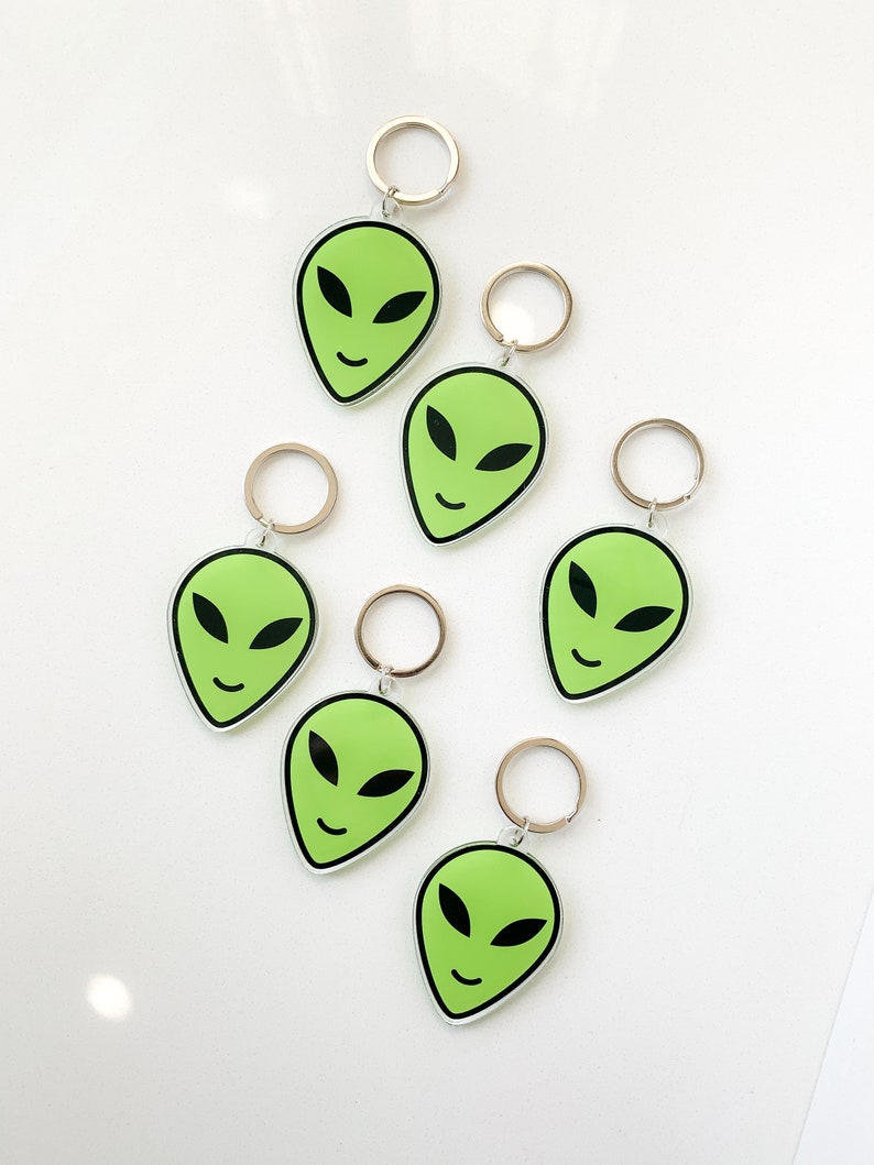 May include: Six green alien face keychains with black eyes and a smile. Each keychain has a silver metal ring.