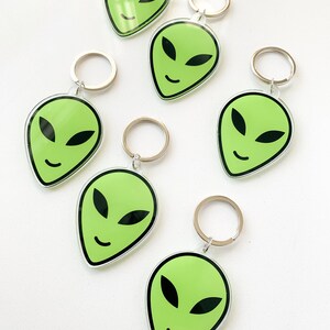 May include: Six green alien face keychains with silver key rings. Each keychain features a green alien face with black eyes and a smile.