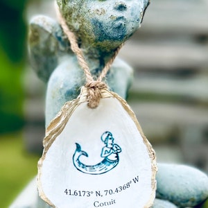 May include: A white oyster shell with a gold rim, featuring a blue mermaid illustration and the text "41.6173° N, 70.4368° W Cotuit".