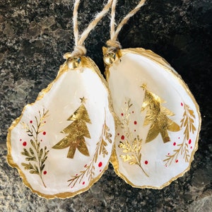 May include: Two oyster shell ornaments featuring a white interior with a gold foil Christmas tree, gold leaf botanical sprigs, and small red dots. The edges are painted gold, and each shell hangs from a natural jute twine loop with a small gold bell.