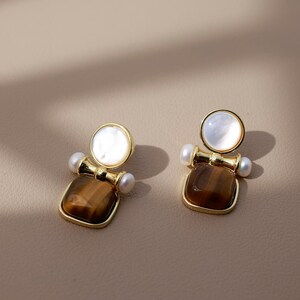 May include: Two gold-coloured earrings. Each has a square, brown tiger's eye base, a small pearl, and a round, white mother-of-pearl top. The earrings are displayed on a neutral surface, with soft shadows.