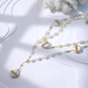 Peut inclure: Un collier à plusieurs rangs avec des perles d'eau douce et des détails en métal doré. Le collier a un design en forme de Y avec plusieurs rangs de perles et de plus grands pendentifs en perles de forme irrégulière enveloppés de fil d'or. Le bijou est présenté sur une surface blanche.