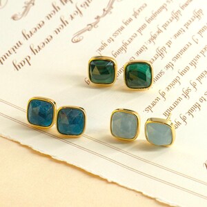 May include: Three pairs of square gemstone stud earrings with gold-coloured settings. The gemstones are blue, green, and light blue. The earrings are displayed on cream-coloured paper with script. Each earring has a gold-coloured bezel.