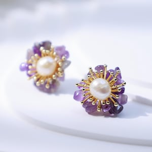 May include: A pair of floral-shaped stud earrings. Each earring features a central white pearl surrounded by small, purple amethyst stones. The earrings have a gold-colored metal setting.