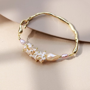 Könnte beinhalten: Ein goldfarbenes Armband mit einem unregelmäßigen, gewellten Metallband. Das Armband ist mit einer Ansammlung kleiner, glänzender weißer und blassrosa Süßwasserperlen in verschiedenen Formen verziert, akzentuiert durch winzige Goldperlen und eine größere.