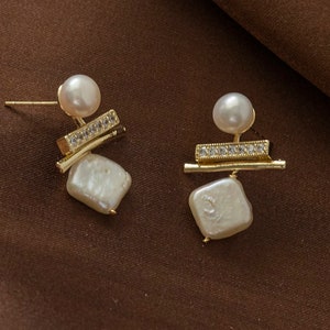 Puede incluir: Un par de pendientes de oro con un diseño geométrico que presenta una perla pequeña y una perla más grande de forma irregular. Los pendientes tienen un estilo minimalista.