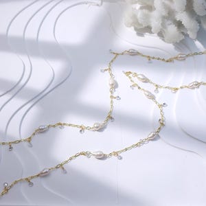 Op de afbeelding: Een delicate gouden ketting met zoetwaterparels en kleine heldere kristallen. De ketting wordt getoond op een witte ondergrond met zachte schaduwen, en een wit koraalachtig object staat op de achtergrond. De parels zijn ovaal van vorm.